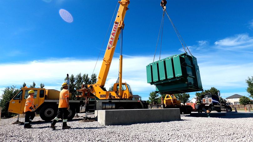 Une grue transporte un grand boîtier électrique vert sous le regard de deux travailleurs portant de l’équipement de sécurité à haute visibilité.