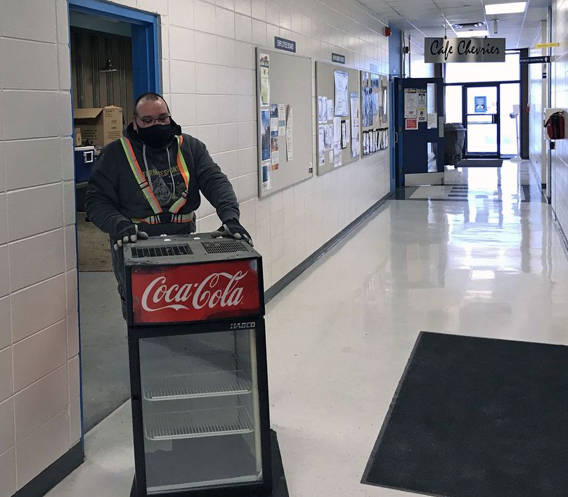 Employee wearing a non-medical mask rolls a small fridge into a hallway in a Manitoba Hydro building.