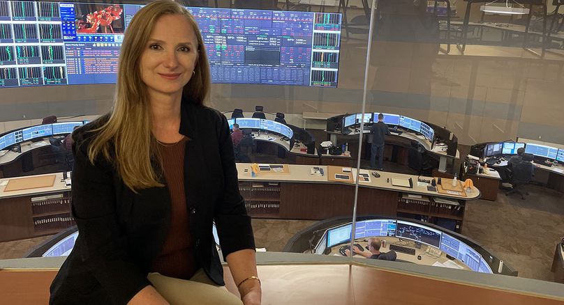 Female employee sitting on desk with large computer screens and work stations in the background.