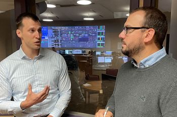 Two male employees talking with large computer screens in the background.