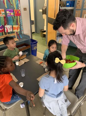 Four children sit around a small table while a man puts vegetables on paper towels beside glasses of milk.