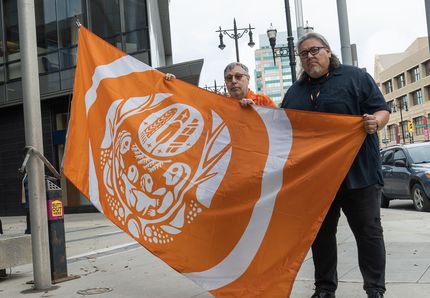 Two Manitoba Hydro employees stand together, holding up the Survivors’ Flag before it goes up the flagpole outside of Manitoba Hydro Place.