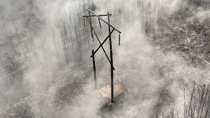 A large transmission line structure made of multiple wooden poles, charred and broken and hanging off power lines amidst a scorched and smoky landscape.