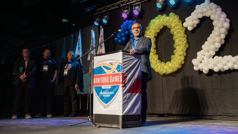 A man speaks at a Manitoba Games podium on a stage decorated with balloons.