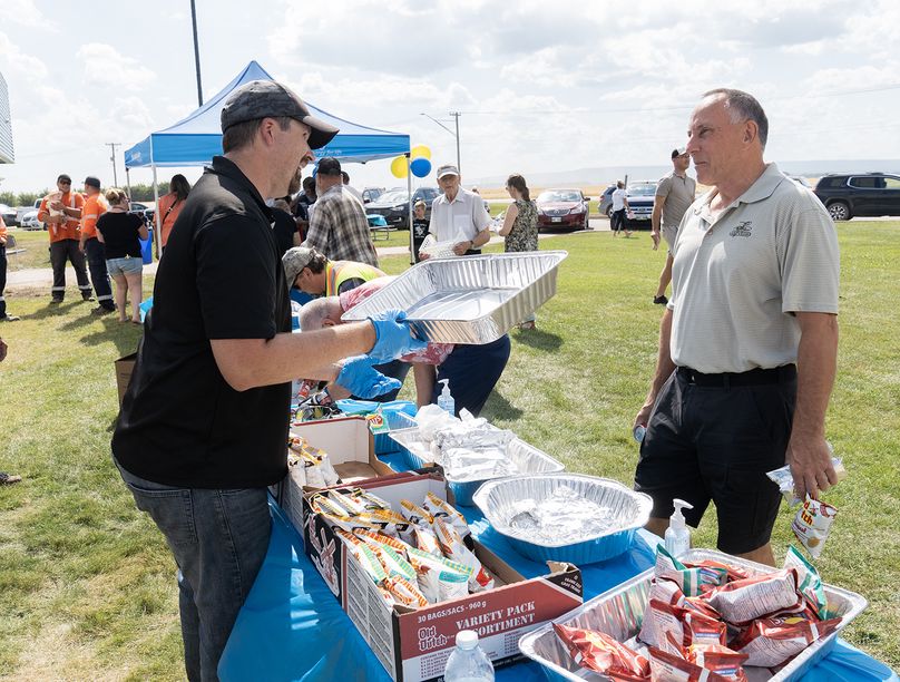 A man serving food chats with a customer over a table of hotdogs and chips.