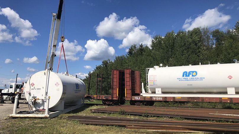 Une grue retire un gros réservoir de carburant métallique d’un wagon de train et le dépose à proximité sur le sol.