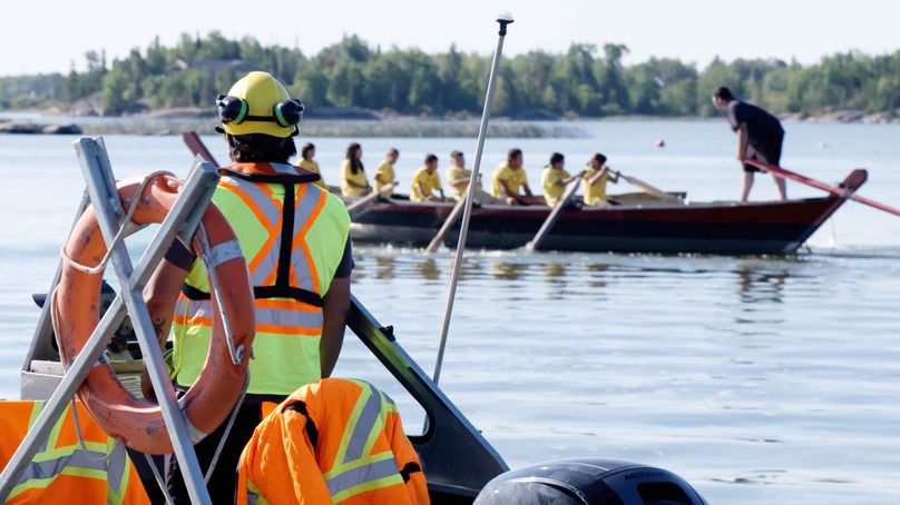 A boat patroller wearing safety equipment watches a team of youths row a large wooden boat.